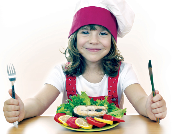 Young girl wearing chef hat and holding fork and knife with plate of food in front of her
