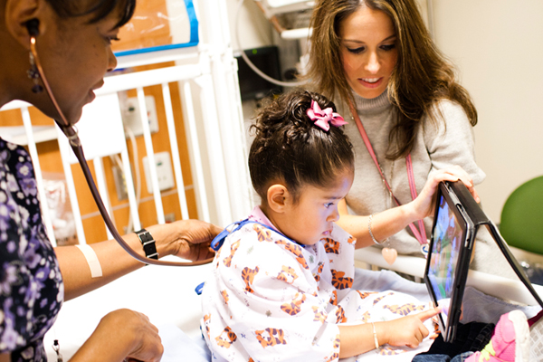 Child life specialist with ipad for patient while nurse listens to her heart