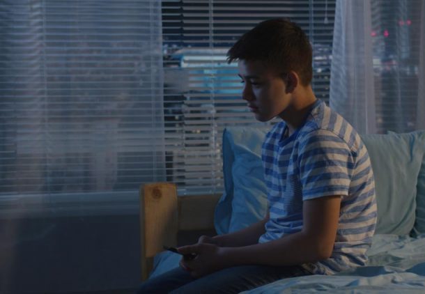 Preteen boy sits on bed in dark room