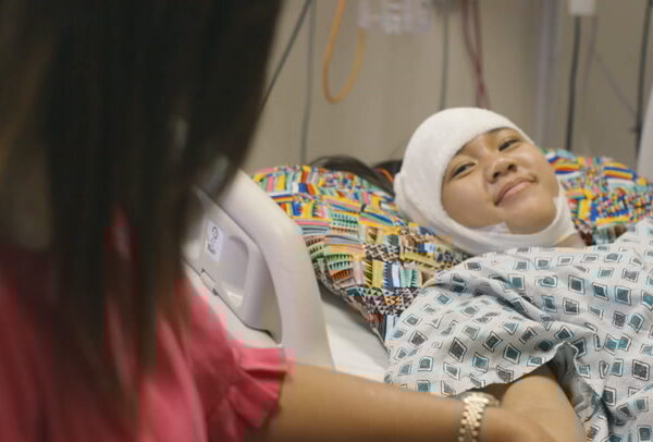 Child in hospital bed with bandaged head, smiling and holding mother's hand.