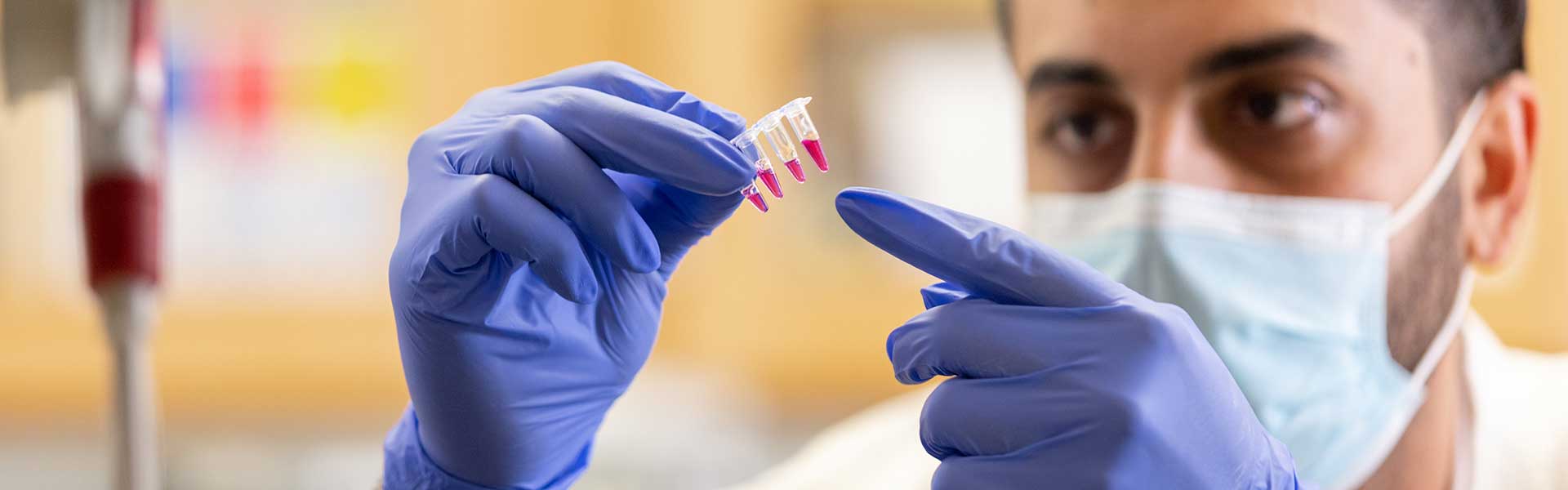 Researcher wearing mask and blue gloves examining pink liquid in a clear tube