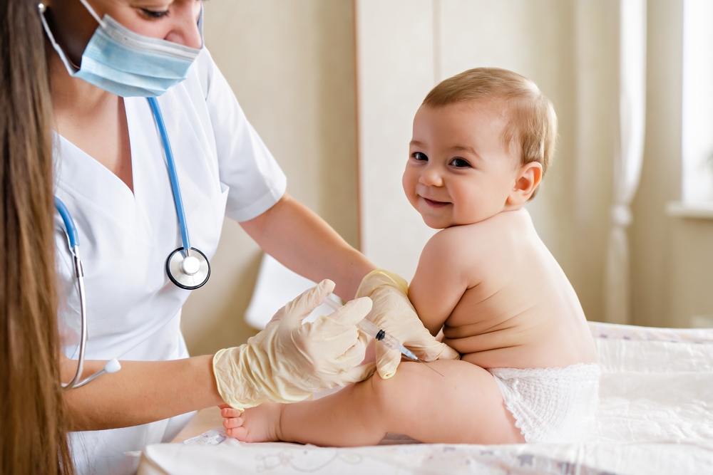 Baby receives an inoculation in the leg while smiling on a table