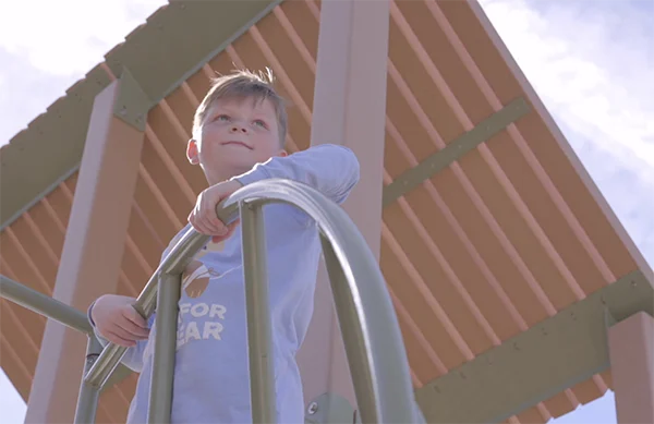 A young boy in a baby blue CHOC shirt looking out at a playground with a smile on his face.