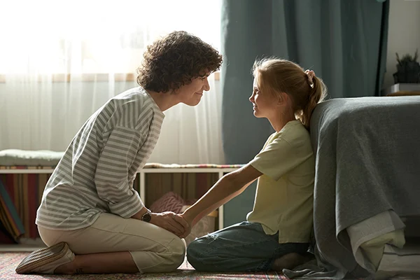 Mother and daughter smiling at each other while holding hands and sitting on the floor in a warmly lit room, showing a moment of connection and support.
