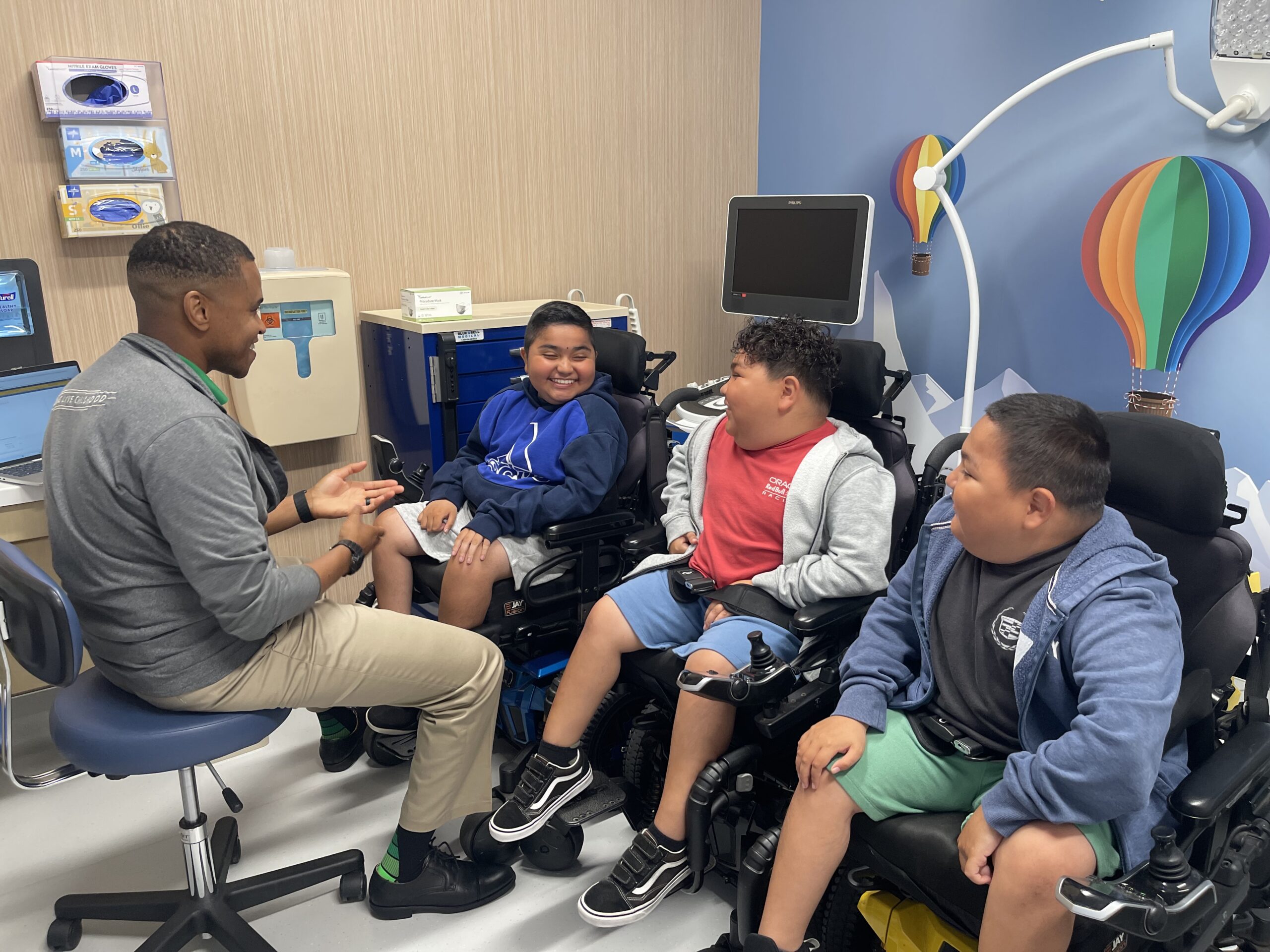 Three young patients laugh and chat with a pediatric neurologist in a welcoming exam room decorated with a colorful hot air balloon.