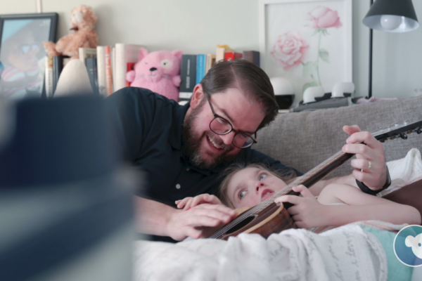 A father playing the ukelele with his daughter in a bright room.