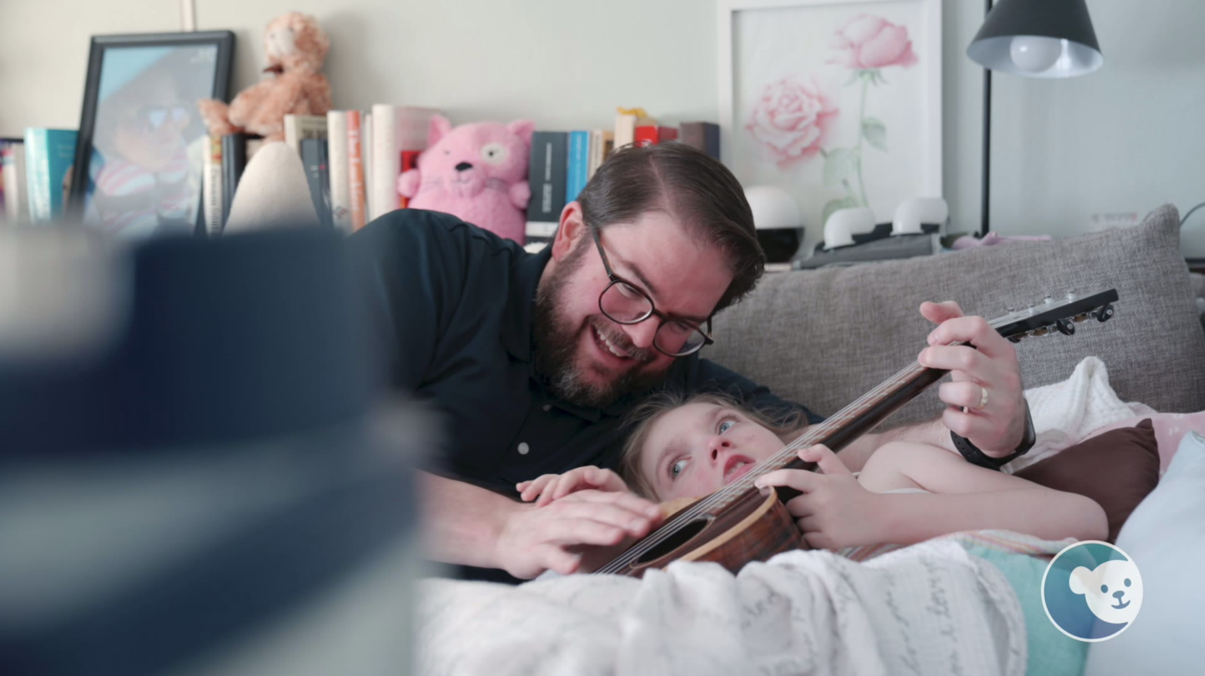 A father playing the ukelele with his daughter in a bright room.
